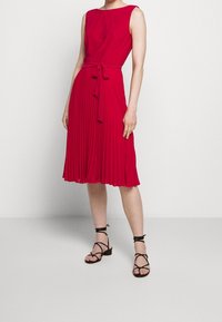 Červené šaty bez rukávů s přiléhavým topem a skladanou sukní, stažené v pase odpovídajícím opaskem. Nosit s černými sandály na vysokém podpatku.