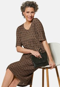 Braun gemustertes Kleid mit kurzen Ärmeln und einem runden Ausschnitt, kombiniert mit einer schwarzen Clutch. Das Kleid zeichnet sich durch ein strukturiertes, geometrisches Design aus.