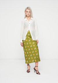 Camisa branca de botões com tecido transparente, emparelhada com uma saia longa verde com um padrão floral e ocular, usada com saltos pretos de tiras.