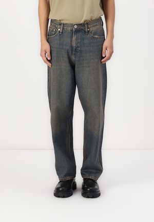 Persoon draagt versleten zwarte loszittende spijkerbroek en zwarte chunky schoenen, staand tegen een effen witte achtergrond.