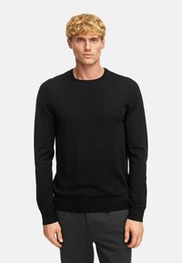 Pull noir à col ras du cou en tissu doux, avec des manches longues et des poignets et un ourlet côtelés, porté sur un pantalon gris foncé.