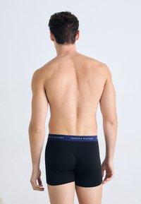 Sorte boksershorts med en marineblå linning som har hvit "TOMMY HILFIGER"-merking. Stoffet ser glatt og tettsittende ut, noe som avslører bakdelen.