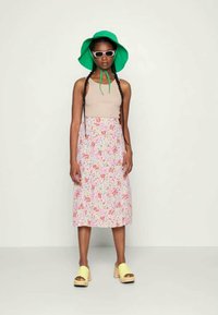 Top sem mangas cor bege, saia midi floral em rosa, branco e verde, chapéu de balde verde, óculos de sol brancos, tamancos amarelos com detalhes em preto.