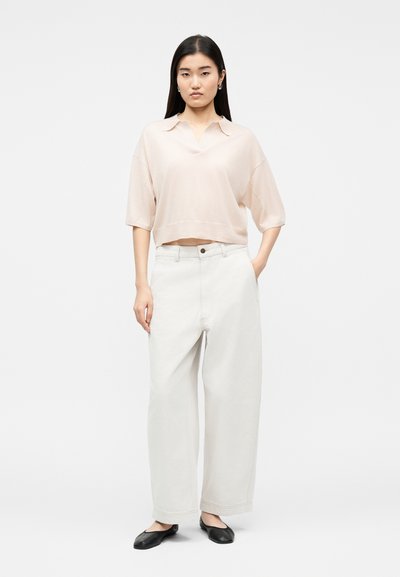 Νεαρή γυναίκα όρθια, φορώντας ανοιχτό ροζ κοντομάνικο τοπ με γιακά, άνετο λευκό παντελόνι φαρδιάς γραμμής και μαύρα επίπεδα παπούτσια, με το χέρι στην τσέπη.