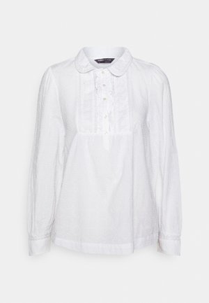 Blouse blanche à manches longues avec col arrondi, bordure en dentelle, patte de boutonnage à l'avant et motif à pois texturé subtil.