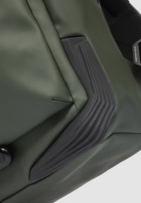 Groene waterdichte tas met een glad oppervlak, voorzien van een zwarte rubberen accenten met getextureerde lijnen en een ronde zwarte knop.