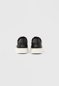 Črni usnjeni športni copati z belo gumijasto podplatom, ravnim petom, oblazinjeno ovratnico in majhnimi logotipskimi poudarki na zadnji strani.