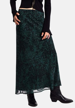 Una falda maxi en verde oscuro con un patrón de estampado animal texturizado. Material translúcido y un diseño fluido, combinada con botas de vaquero negras.