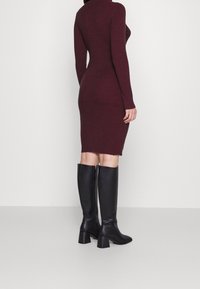 Burgundská pletená šaty se žebrovaným vzorem, dlouhými rukávy aúzkým střihem, délka po kolena. V kombinaci s vysokými černými koženými botami na vysokém podpatku.