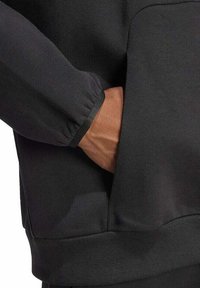 Manga de sudadera negra con puño elástico, presentando una mano en un bolsillo frontal. El material parece suave y liso, con un diseño de corte relajado.