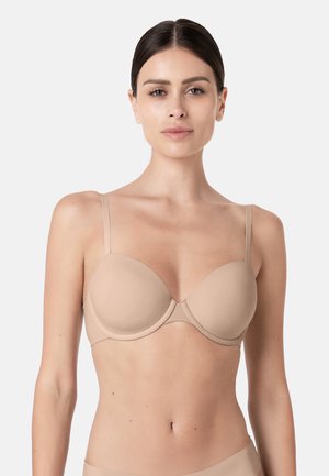 Donna con capelli scuri che indossa un reggiseno imbottito beige con spalline sottili, rivolta in avanti su uno sfondo chiaro e uniforme.