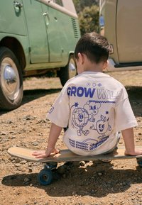 Witte t-shirt met blauwe grafische ontwerpen van lachende figuren en de tekst "GROW WILD" op de achterkant. Kind zittend op een houten skateboard.