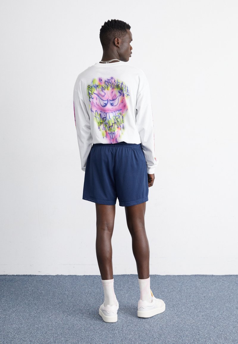 Camisa blanca de manga larga con un gráfico multicolor en la espalda, combinada con pantalones cortos azul oscuro, zapatillas blancas y calcetines claros.