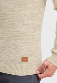 Vaaleanbeige neulepullover, jossa on hienovarainen rakenne, ribbaatut hihansuut ja helma, sekä nahkaleima, jossa on brändin nimi alareunassa.