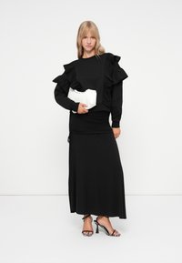 Hanorac negru cu mâneci volan, asortat cu o fustă neagră plisată. Modelul ține o geantă albă cu cusături decorative și poartă pantofi cu toc subțire.