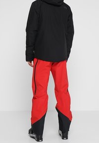 Rote wasserdichte Schneehosen mit schwarzen Akzenten, ausgestattet mit Seitenreißverschlüssen und einer lockeren Passform, entworfen für den Wintersport und Outdoor-Aktivitäten.