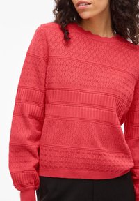 Vrouw met een rood gebreide trui met diamant- en ribbeltjespatronen, gecombineerd met een zwarte broek, zichtbaar van borst tot taille.