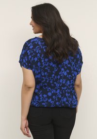 Blommig marinblå blus med korta ärmar, med blå blommiga mönster. Materialet verkar lätt och har en lös passform. Stylar med svarta byxor.