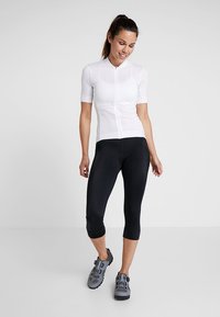 Jersey de ciclismo branco de manga curta com fecho à frente, combinado com leggings capri pretas. Sapatos de ciclismo cinzentos com tiras são visíveis.