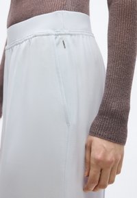 Közelkép egy személyről, aki barna bordázott hosszú ujjú felsőt és fehér, oldalzsebes, gumis derekú nadrágot visel, keze az oldalán pihen.