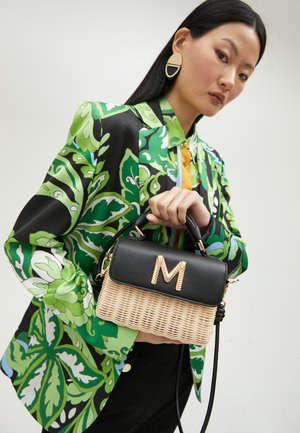 Bolso de paja negro y trenzado con un asa superior, logotipo dorado "M" y una correa desmontable, sostenido por un modelo que lleva un vibrante blazer floral.