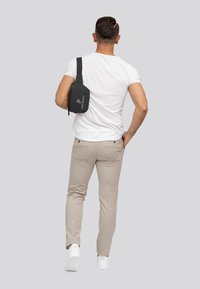 Zwarte schoudertas met een gladde afwerking, met het logo "parcatistic," gedragen door een persoon in een wit shirt en lichtgekleurde broek.