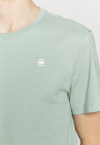 T-shirt in cotone verde chiaro con colletto rotondo e maniche corte. Presenta un piccolo logo bianco sul petto. Texture liscia; design casual.