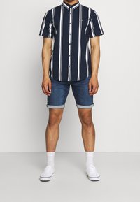 Marinblå kortärmad skjorta med vita vertikala ränder, ståkrage, knäppning med knappar, bärs med denimshorts och vita sneakers samt strumpor.