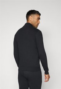 Casaco preto com fecho zip em tecido suave, apresentando uma gola alta e punhos ajustados. A vista traseira mostra um design elegante sem padrões ou acessórios visíveis.