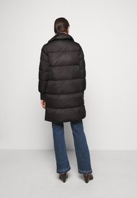 Svart pufferjacka med hög krage, med horisontell quiltning och långa ärmar, kombinerad med utsvängda blå jeans och spetsiga bruna skor.