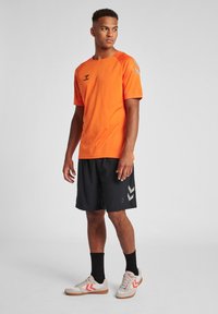 Oranje sportshirt met korte mouwen, gecombineerd met zwarte shorts met grijze chevronaccenten. Draag met beige sneakers en zwarte sokken.