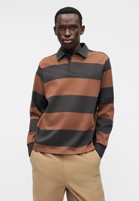 Καφέ και μαύρο ριγέ μακρυμάνικο πόλο με γιακά, που διαθέτει ανοιχτό πατιλέτα και μαλακή υφή, σε συνδυασμό με μπεζ παντελόνι από κορντόνι.