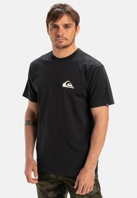 Black cotton t-shirt featuring a small white logo on the left chest. Crew neck design, short sleeves, paired with camouflage pants.