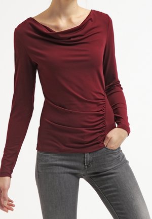 Vrouw draagt een aansluitende bordeauxrode top met lange mouwen, een gedrapeerde halslijn en gerimpelde details, gecombineerd met een grijze spijkerbroek, hand in de zak.