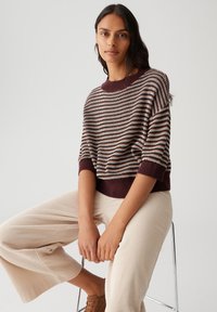 Woman sitting on a stool wearing a striped brown and white knit sweater, beige wide-leg pants, and brown shoes against a plain background.