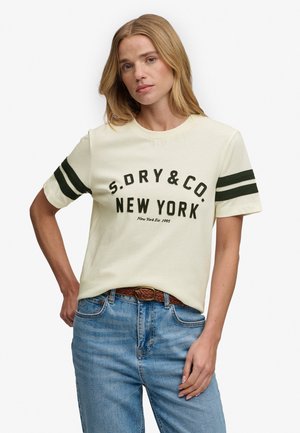 Kobieta nosząca kremowy t-shirt z czarnym napisem „S.DRY & CO. NEW YORK” i podwójnymi czarnymi paskami na rękawach, w połączeniu z niebieskimi dżinsami i brązowym paskiem.