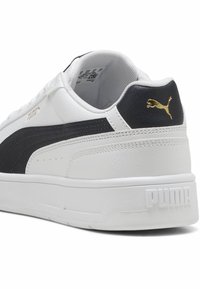 Hvide sneakers med en tekstureret læderoverflade, der har en sort sideløsning og guld logo accenter på hælen. Flad gummisål.
