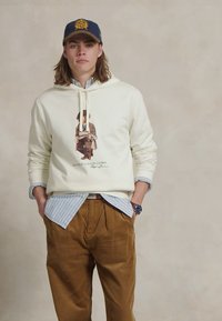 Crème hoodie met een frontafbeelding, over een lichtblauw gestreept shirt. Bruine corduroy broek, marineblauwe pet met gouden embleem. Casual kleding.
