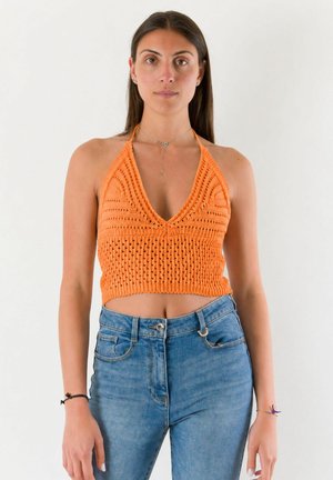 Donna con top arancione all'uncinetto a collo halter e jeans blu, in piedi davanti a uno sfondo chiaro e uniforme.