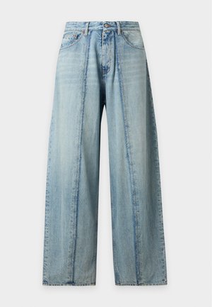 Mann trägt ein schlichtes weißes oversized T-Shirt und locker sitzende hellblaue Jeans und steht vor einem weißen Hintergrund.