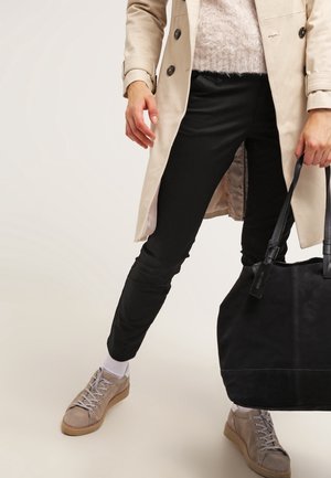 Person iført beige trenchcoat, sorte bukser, beige sneakers, der holder en stor sort håndtaske mod en ensfarvet lys baggrund.