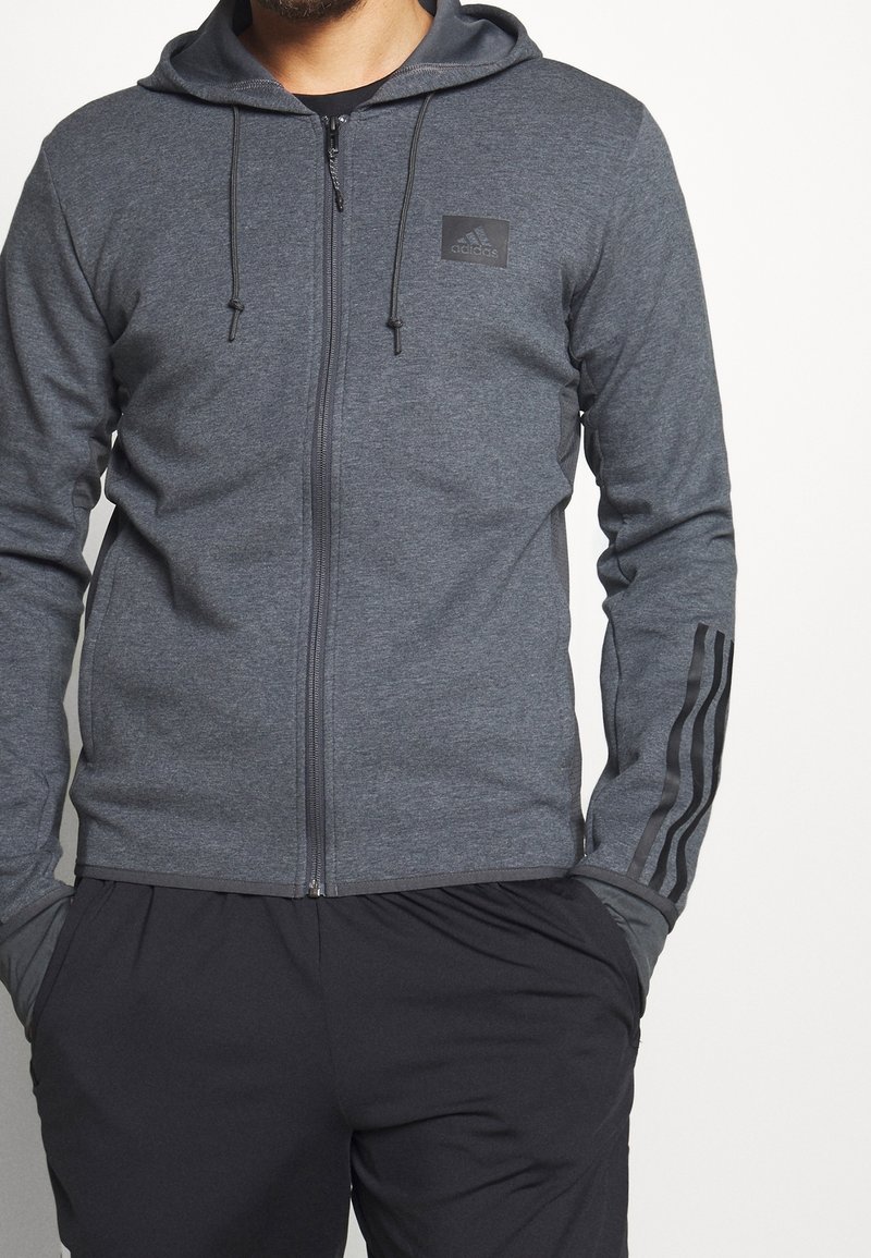 Grauer Sweatshirt mit Reißverschluss, Kapuze und schwarzem Logo. Verfügt über dunkle Streifenakzente an den Ärmeln und einen glatten, strukturierten Stoff.