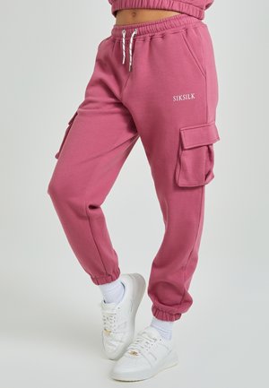 Personne portant un pantalon cargo rose avec des ourlets élastiques, des baskets blanches et des chaussettes blanches sur un fond clair uni.