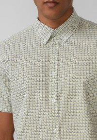 Kurzarmhemd aus einem hellgelben Stoff, mit einem geometrischen Muster in Blau und Lila, mit einem Button-down-Kragen und kleinen Knöpfen.