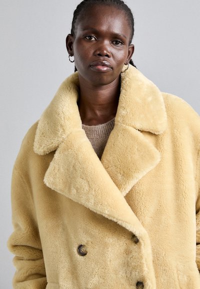 Gelber Kunstpelzmantel mit Doppelknopf-Design, großem Kragen und zwei Knöpfen. Getragen über einem beigen Pullover mit weicher Textur.
