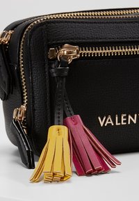 Černá kožená taška s texturou a zlatými kovovými prvky. Obsahuje barevné třásně v žluté a burgundské barvě. S termo uzavíratelnými přihrádkami pro úložný prostor.