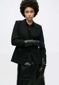 Μαύρο σακάκι με ραφή, με χρυσές λεπτομέρειες, συνδυασμένο με μαύρο πλεκτό πουκάμισο και δερμάτινα γάντια. Λεπτό υφή και εφαρμοστή σχεδίαση.