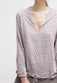 Blouse à manches longues en tissu gris clair avec un décolleté en V et un motif ovale répétitif en rose et crème. Dispose d'un cordon de serrage au bas.