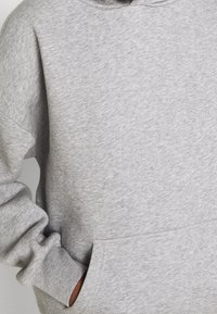 Personne portant un sweat-shirt gris clair avec la main partiellement dans la poche avant et le poignet retroussé visible.