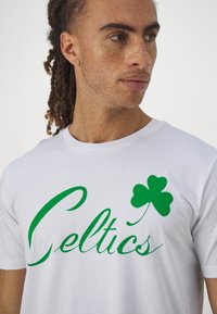 Biała bawełniana koszulka z krótkim rękawem z zielonym napisem "Celtics" oraz wzorem zielonego koniczka. Posiada okrągły dekolt. Gładka tekstura.